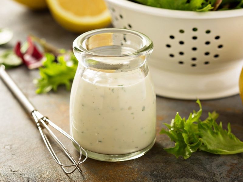 Joghurt-Dressing vor Sieb mit Salat auf grauer Oberfläche. Daneben liegen ein Schneebesen, einige Salatblätter und angeschnittene Zitronen.