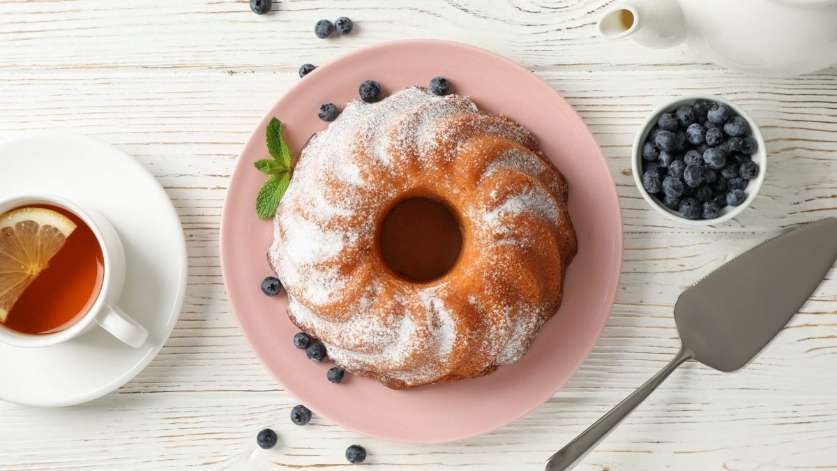 Gugelhupf mit Puderzucker auf rosa Teller. Dieser steht auf einem weißen Holztisch, umgeben von Heidelbeeren und einer Tasse Tee mit Zitrone.
