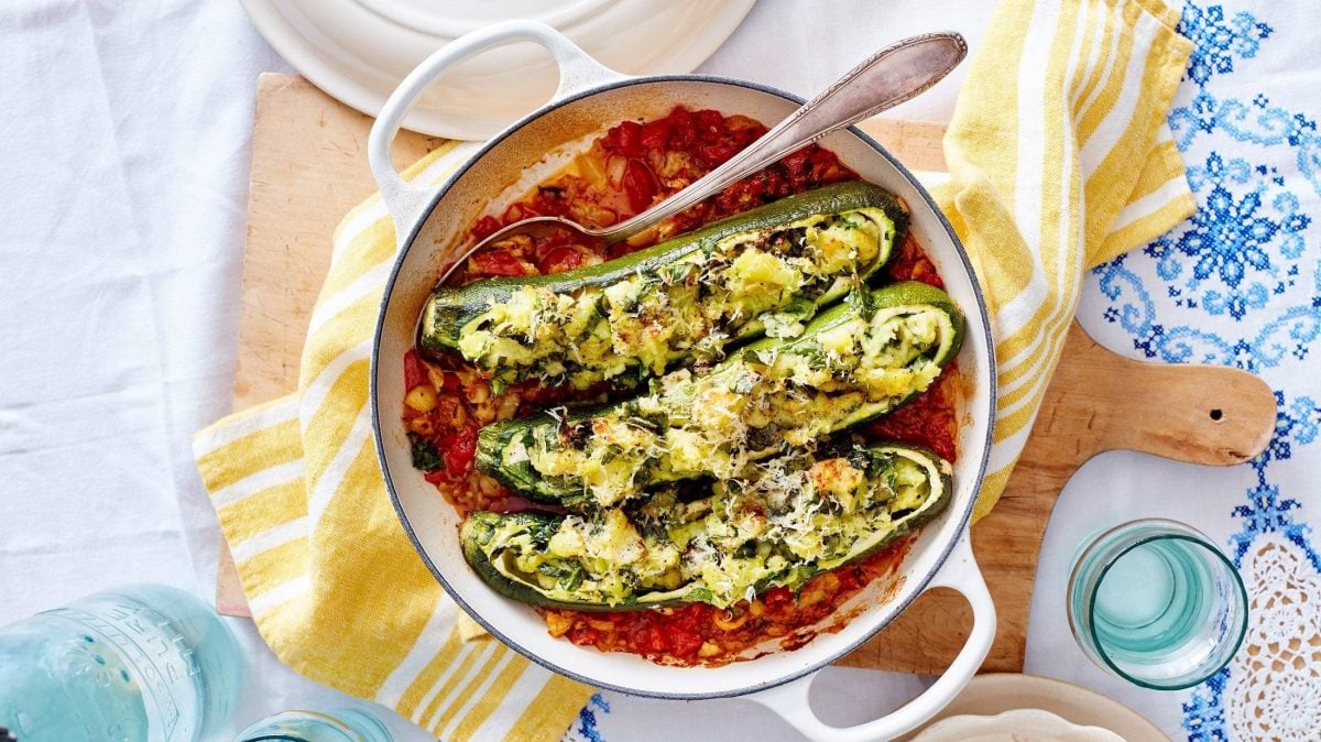 Drei Zucchini-Kartoffel-Schiffchen liegen in der Sauce in einem Bräter. Dieser steht auf einem Holzbrett auf dem Tisch.