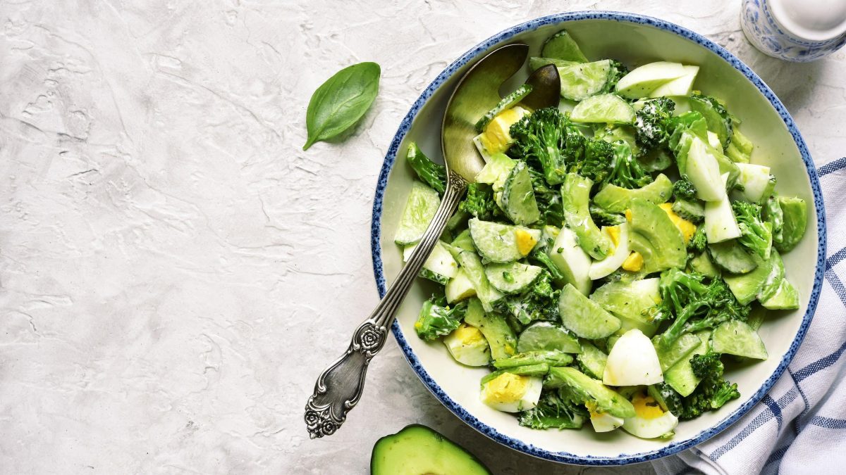 Saftiger Avocadosalat mit Brokkoli, Gurke und Ei mit einer feinen Dill-Sauce und Basilikumblättern in einer hellen Schale auf einem Steintisch serviert.