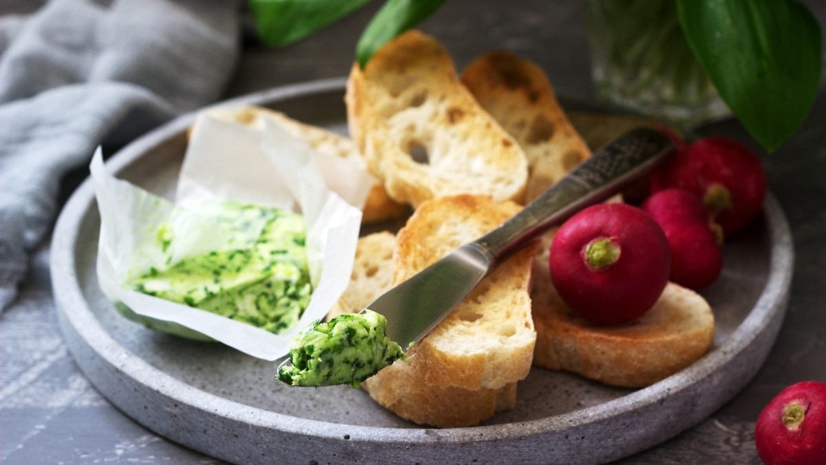 Bärlauch-Butter in Papier auf grauem Teller mit Baguette-Scheiben und Radieschen. Auf dem Brot ein Messer mit Bärlauch-Butter- Im Hintergrund graues Tuch. Frontalansicht.