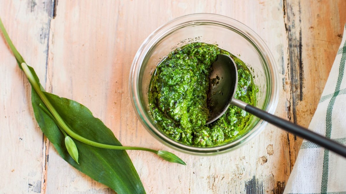 Bärlauch-Pesto in einem Glas mit silbernem Löffel. Daneben frischer Bärlauch. Angerichtet auch Holzbrett. Links am Bildrand blau-weißes Leinentuch. Aufnahme aus der Vogelperspektive.