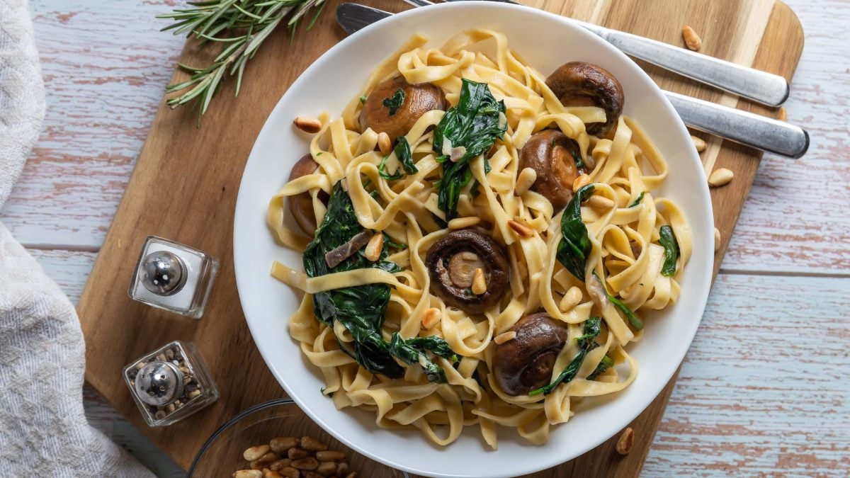 Ein Teller Bärlauch-Spaghetti mit Champignons und Pinienkernen auf einem Brett in der Draufsicht, daneben Gewürzstreuer und Besteck.