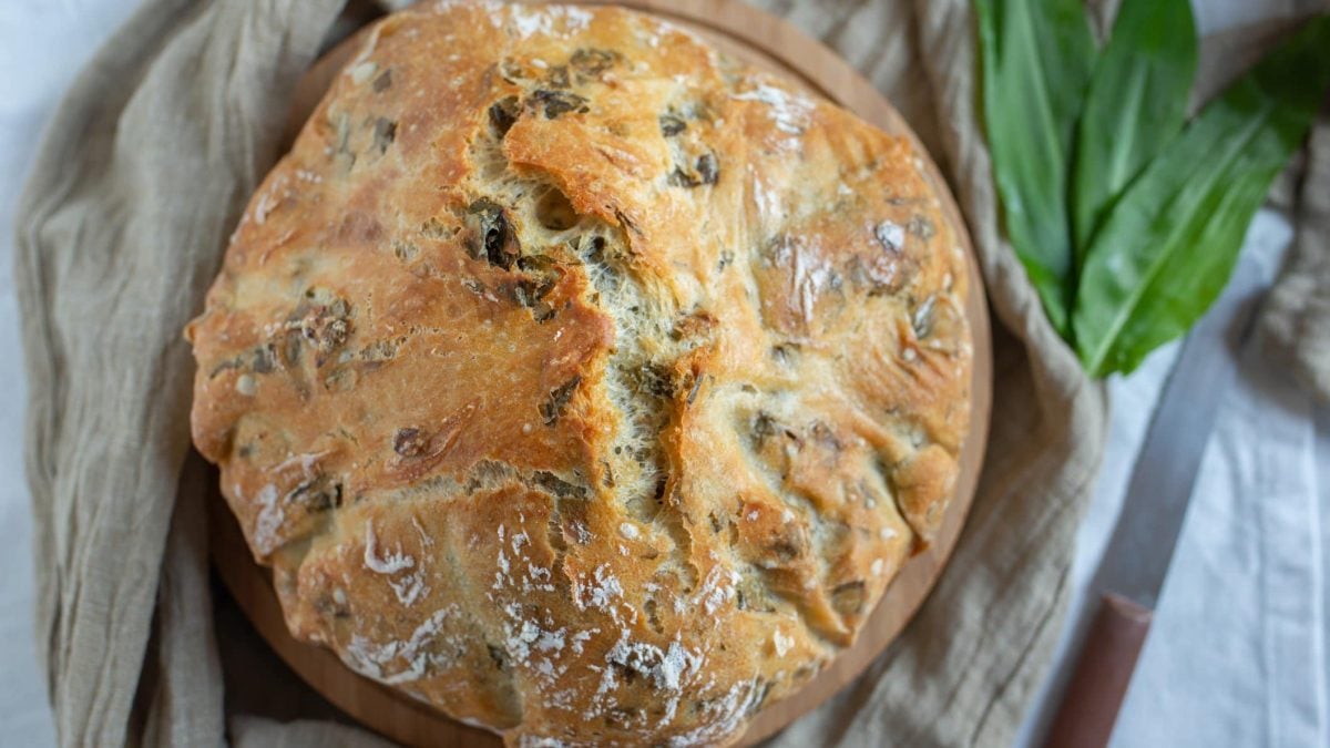 Ein Rundes Bärlauchbrot mit Nüssen in der Draufsicht, daneben frischer Bärlauch und ein Messer.