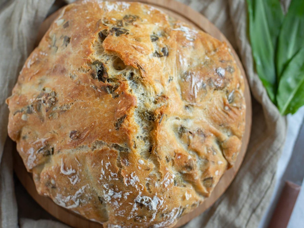 Ein Rundes Bärlauchbrot mit Nüssen in der Draufsicht, daneben frischer Bärlauch und ein Messer.