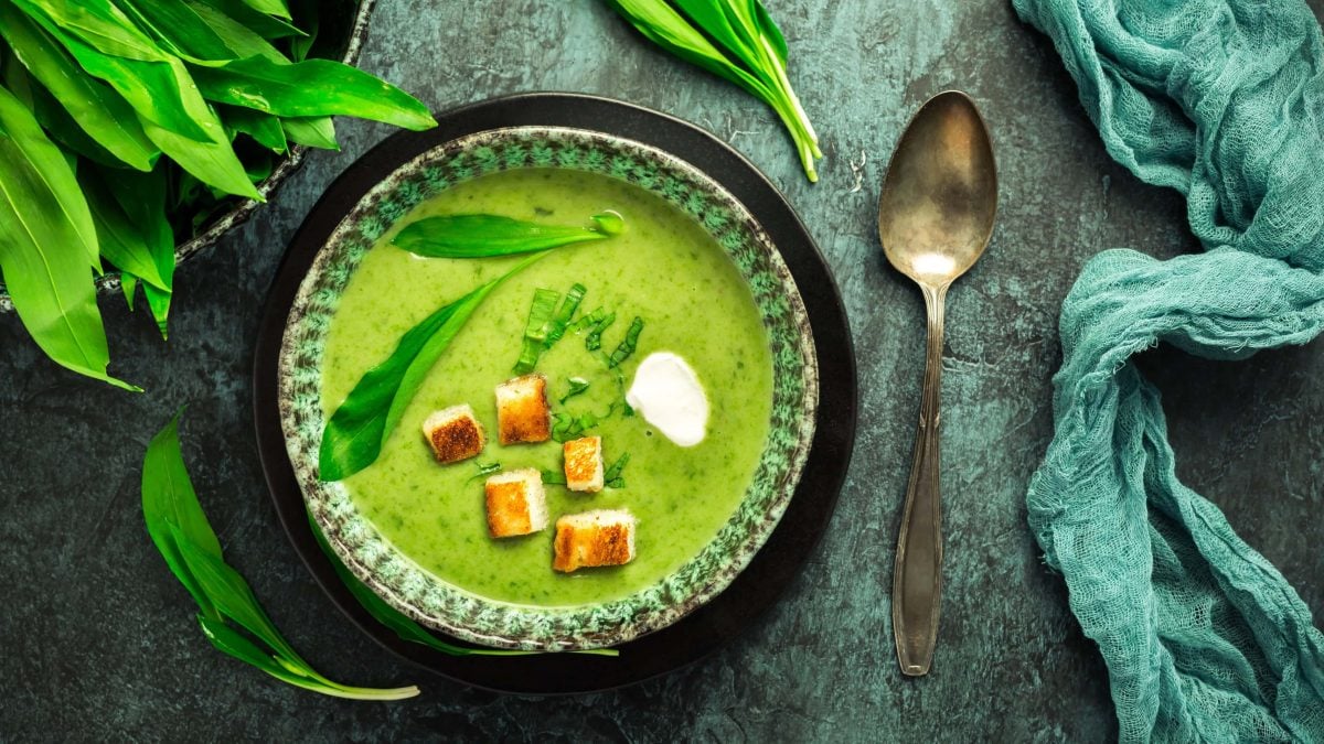 Ein Teller Bärlauchsuppe mit Croutons in der Draufsicht, daneben ein Löffel, Geschirrtuch und frischer Bärlauch.