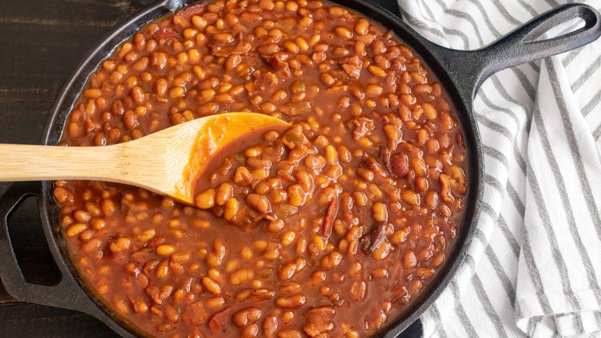 Selbst gemachte Baked Beans Heinz Beanz in schwarzer Guss-Pfanne auf dunklem Untergrund mit Holzlöffel. Daneben gestreiftes Stofftuch.