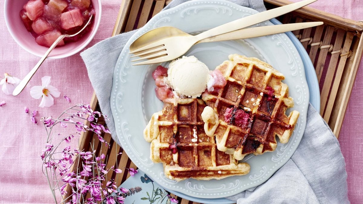 Belgische Rhabarber-Waffeln mit fruchtigem selbstgemachten Rhabarberkompott und einer Kugel Eis auf einem hellen Teller auf einem mit rosaner Tischdecke verzierten Tisch.