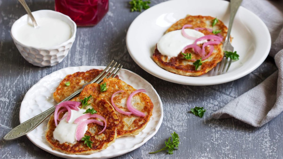 Saftige Boxty-Pancakes, bestehend aus Pfannkuchenteig mit Sahne und geriebener Kartoffel, serviert mit Schmand, eingelegten Zwiebeln in Essig und frischer Petersilie auf hellen Tellern auf hölzernem Untergrund.
