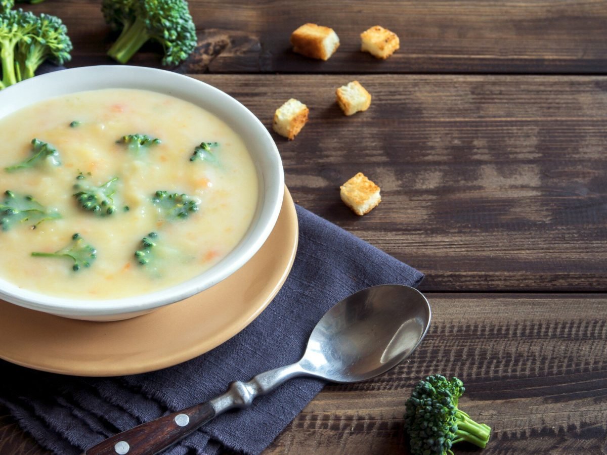 Brokkoli Cheddar Suppe auf einem weißen Teller. Daneben verschiedene Zutaten, Küchentücher und ein Löffel. Das Ganze auf einem hölzernen Untergrund.