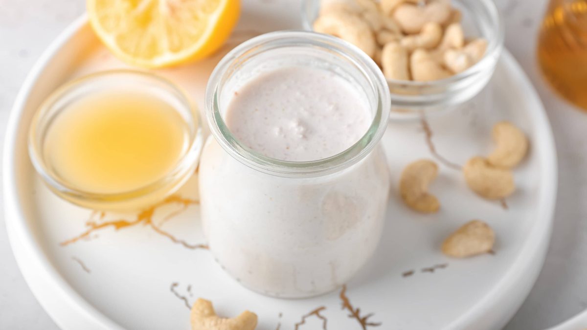 Ein Glas Cashewsahne auf einem weißen Tablett, daneben eine Schale Cashewkerne und eine Schale Zitronensaft.