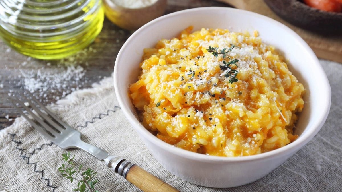 Cheddar Risotto in einer weißen Schüssel. Daneben einige Zutaten sowie eine Gabel. Das Ganze auf einem Küchentuch auf einem hölzernen Untergrund.