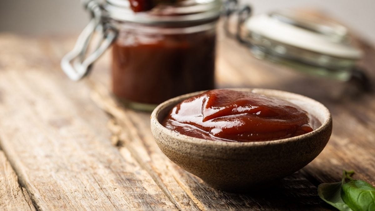 Dr. Pepper Barbecue-Sauce in brauner Schale auf Holztisch. Im Hintergrund Sauce im Einweckglas. Frontalansicht.