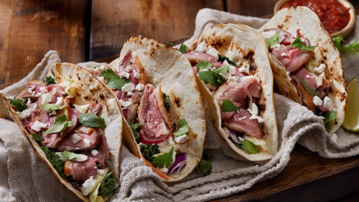 Vier mexikanische Entenbrust-Tacos auf beigem Tuch auf dunklem Untergrund. Draufsicht.