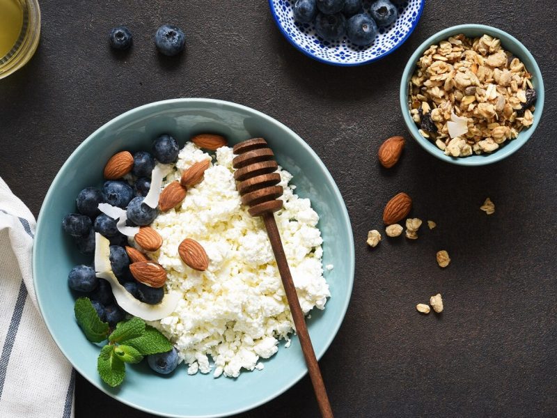 Unsere Hüttenkäse-Frühstücks-Bowl steht angerichtet in einer Schüssel auf dem Tisch. Daneben stehen in kleinen Schälchen Honig, Blaubeeren und Nüsse.
