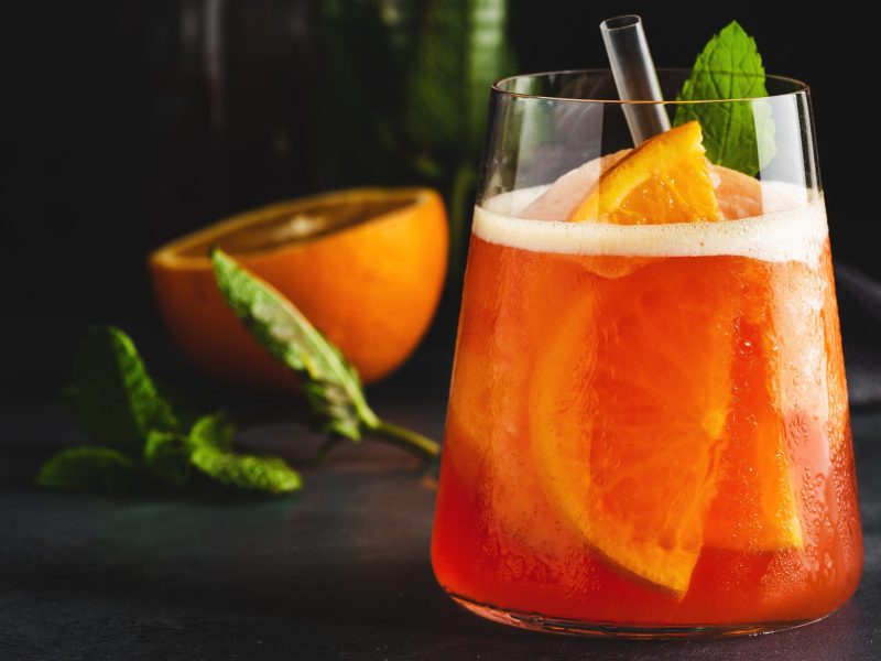 Gin and Sin Gin Cocktail mit Orangen und Minzblatt im Glas auf schwarzem Tisch. Im Hntergrund halbe Orange und Minze. Frontalansicht.