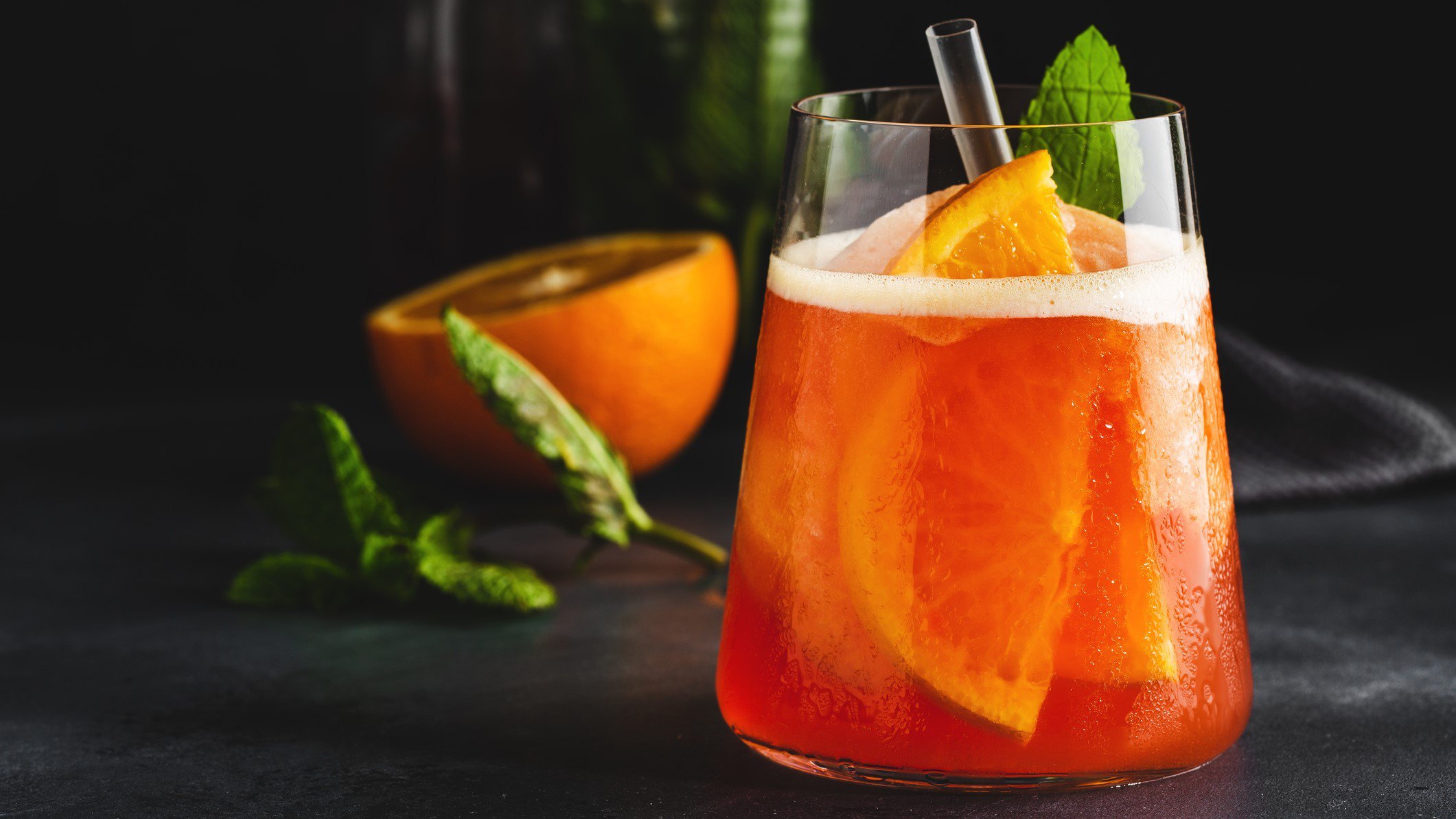 Gin and Sin Gin Cocktail mit Orangen und Minzblatt im Glas auf schwarzem Tisch. Im Hntergrund halbe Orange und Minze. Frontalansicht.