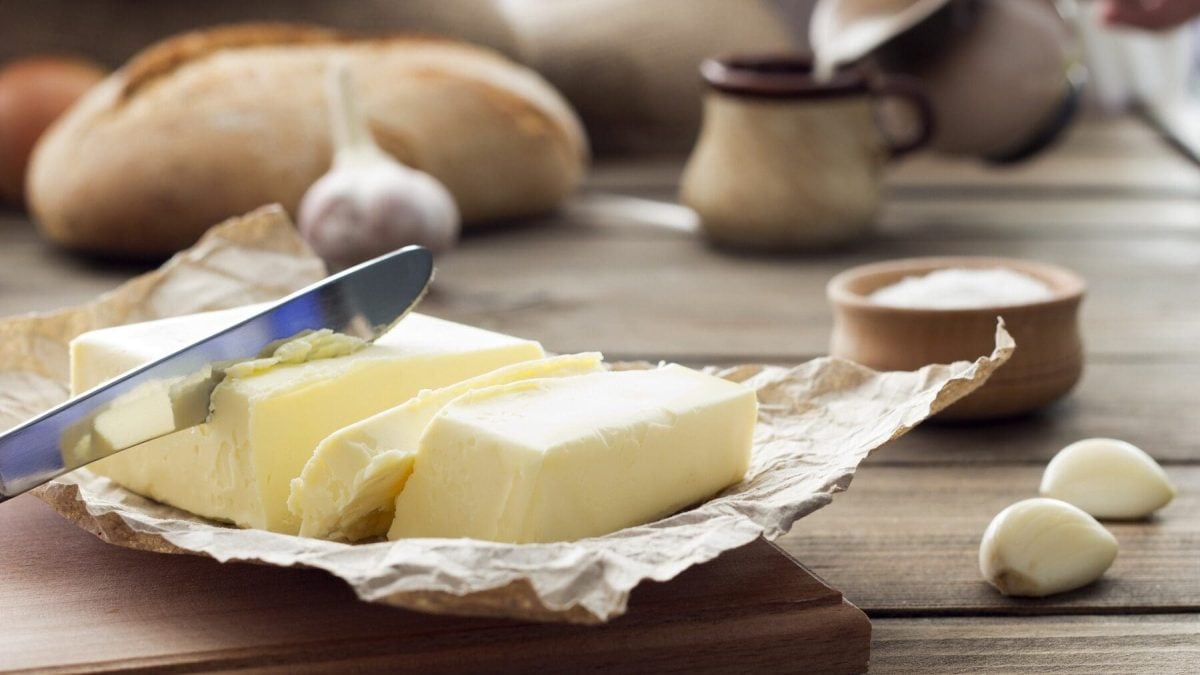 Auf einem Holztisch liegen Butter im Packpapier, zwei Knoblacuzehen, Eier, Brot und eine Schale mit einer weißen Masse drin.