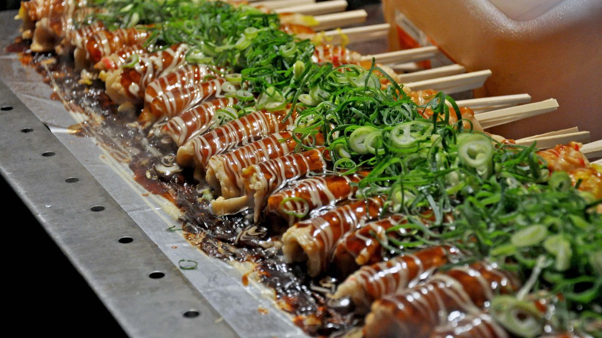 Hashimaki auf Gasgrill aus Metall an Streetfood-Stand in Japan. Im Hintergrund Kanister mit Öl. Frontalansicht.