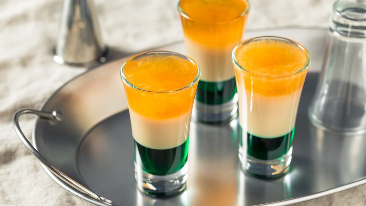 Drei selbstgemachte Irish Flag Shots auf metallsicher Servierplatte. Umgedrehtes Shotglas daneben. Im Hintergrund Barmaß. Frontalaufnahme.