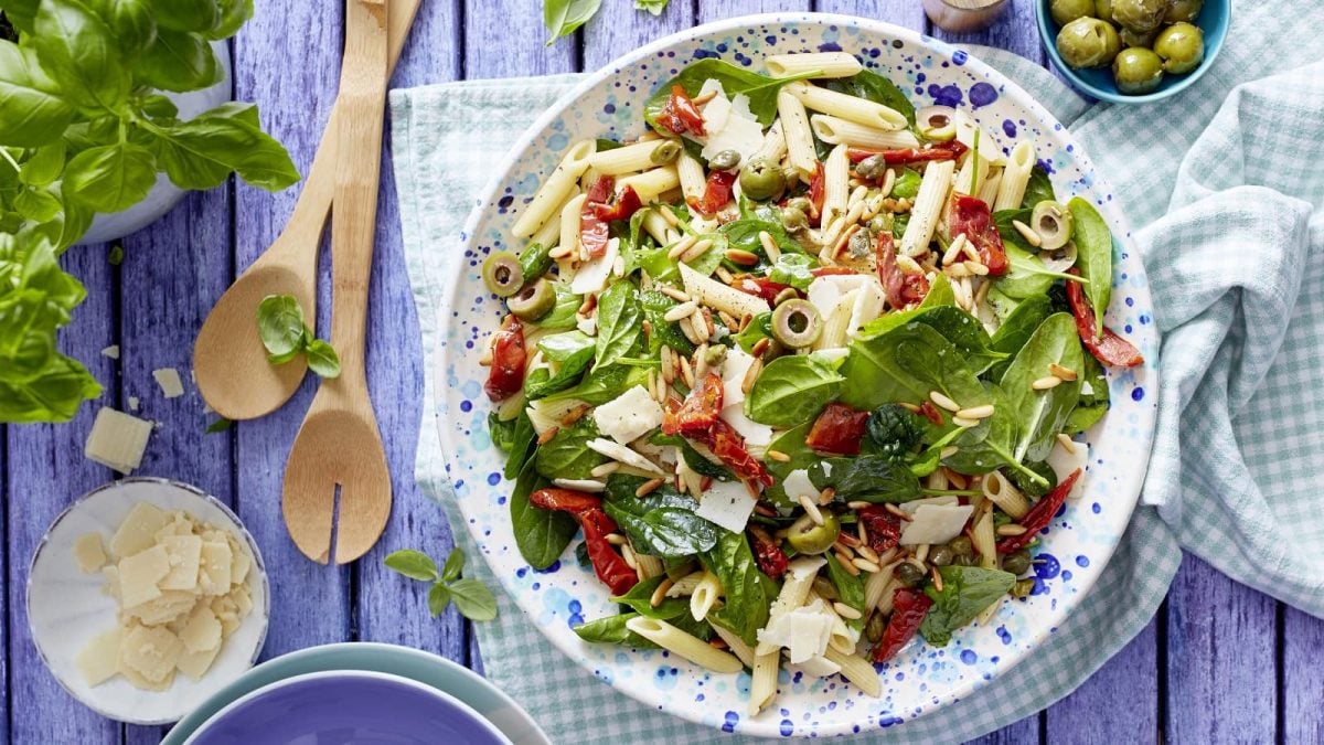 Italienischer Nudelsalat mit Oliven und frischem Basilikum mit Parmesan auf einem Teller auf einem gedeckten Tisch neben zwei Kochlöffeln