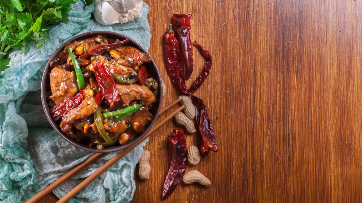 Kung Pao Chicken in schwarzer Schüssel auf blauem Tuch auf Holztisch. Daneben getrocknete Chilis, Essstäbchen, Knoblauch und Koriander. Aufnahme aus der Vogelperspektive.