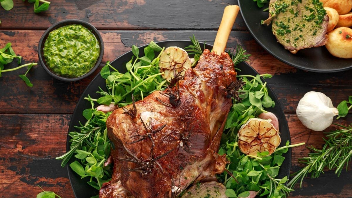Geschmorter Lammkeule mit Minz-Sauce auf Wildkräutern auf schwarzem Teller und Holztisch. Daneben Schale mit Minz-Sauce. Oben Teller mit Röst-Kartoffeln und Bratenscheiben mit Sauce. Rosmarin, Knoblauch und Kräuter als Deko verteilt. Aufnahme aus der Vogelperspektive.