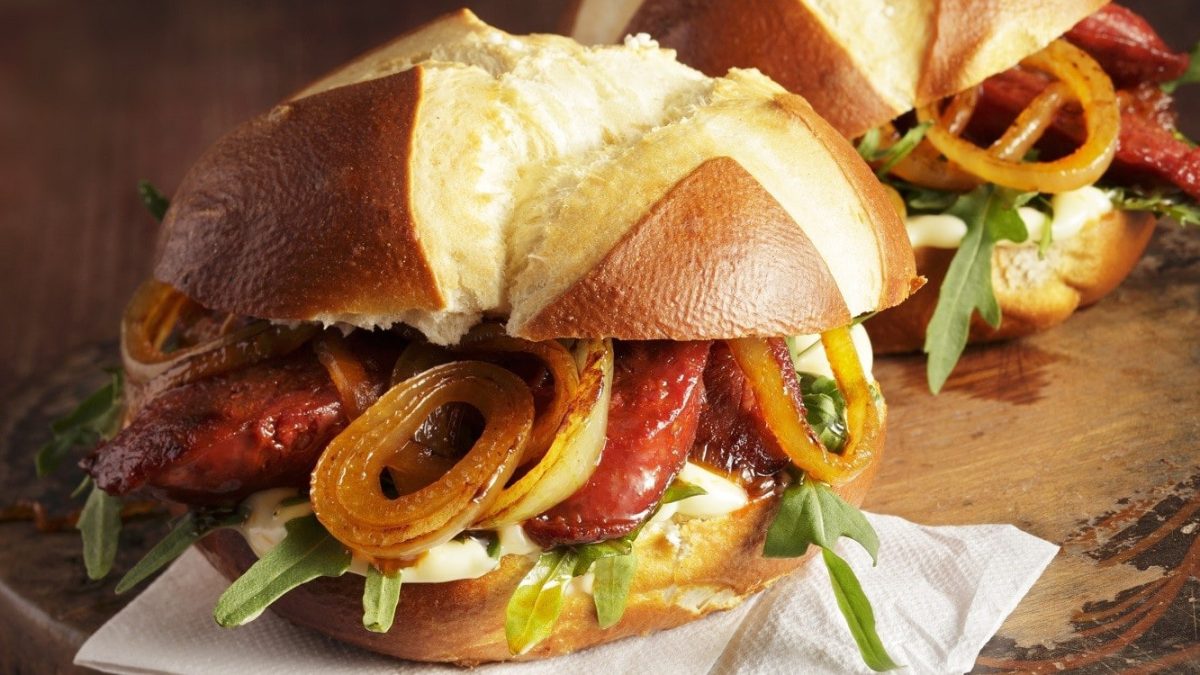 Ein Laugenbrötchen mit Chorizo und Röstzwiebeln, garniert mit frischem Rucola und einem feinen Dip, serviert auf einem Holzbrett