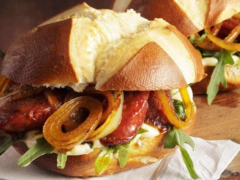 Ein Laugenbrötchen mit Chorizo und Röstzwiebeln, garniert mit frischem Rucola und einem feinen Dip, serviert auf einem Holzbrett