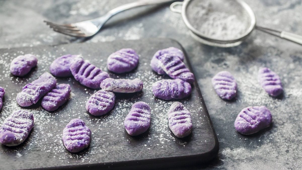 Aus der Vogelperspektive: Auf einer schwarzen Schieferplatte liegen Lila-Kartoffel-Gnocchi, eine Gabel und ein kleines Sieb mit einem weißen Pulver. Es handelt sich um Mehl-