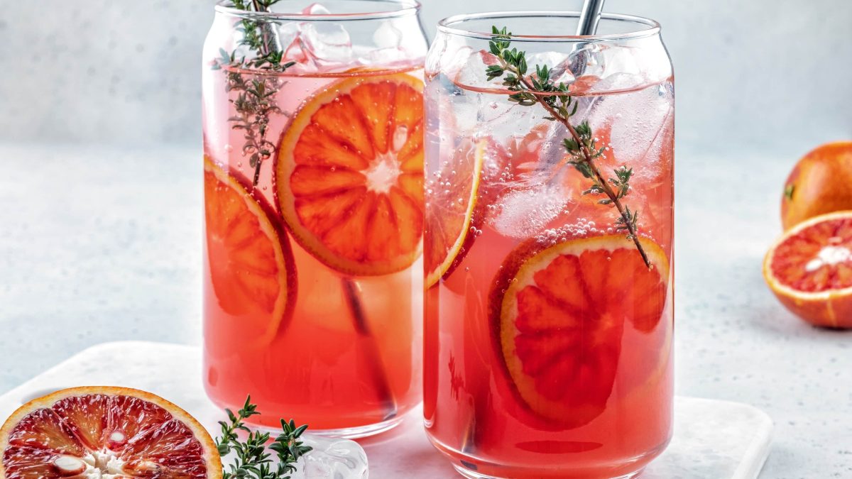 Make it Count Campari Cocktail mit Blutorange und Eiswürfeln und Thymianzweig im Glas auf Glastisch vor hellem Hintergrund. Blutorangenscheiben und Thymian als Dekoration im Bild. Frontaaufnahme.