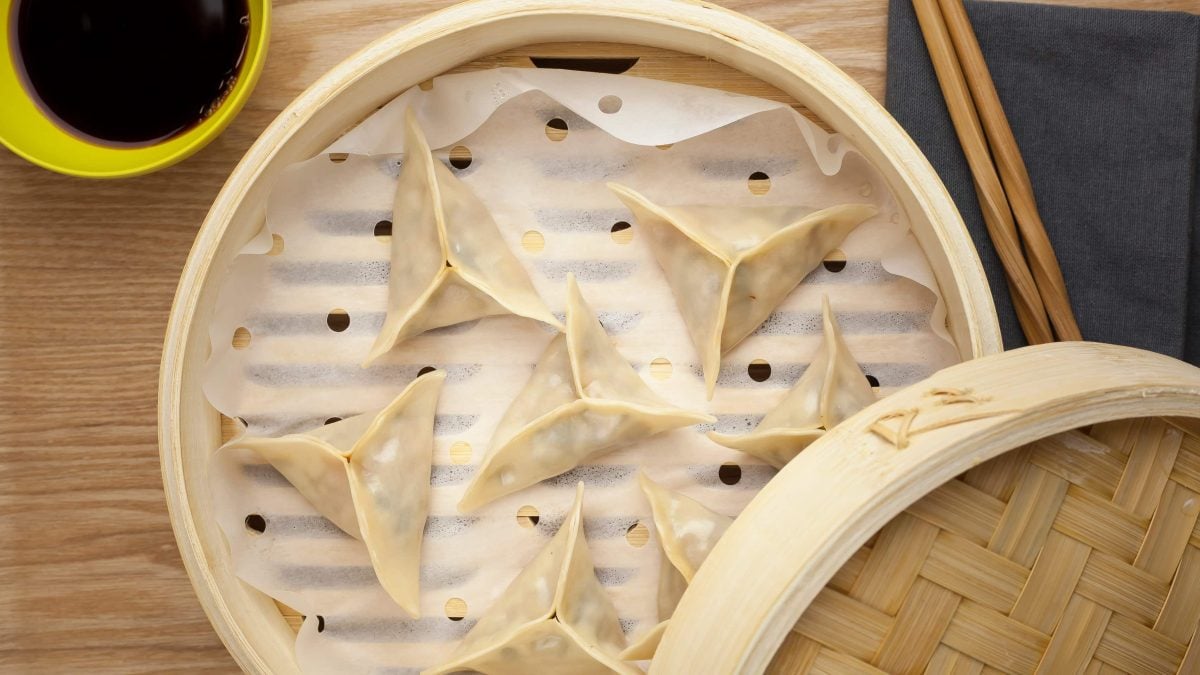 Vegetarische Mandu mit selbst gemachtem Kimchi gefüllt im Bambus-Dämpfer auf Holztisch. Daneben gelbe Schale mit dunkler Sauce. Rechts Essstäbchen. Aufnahme aus der Vogelperspektive.