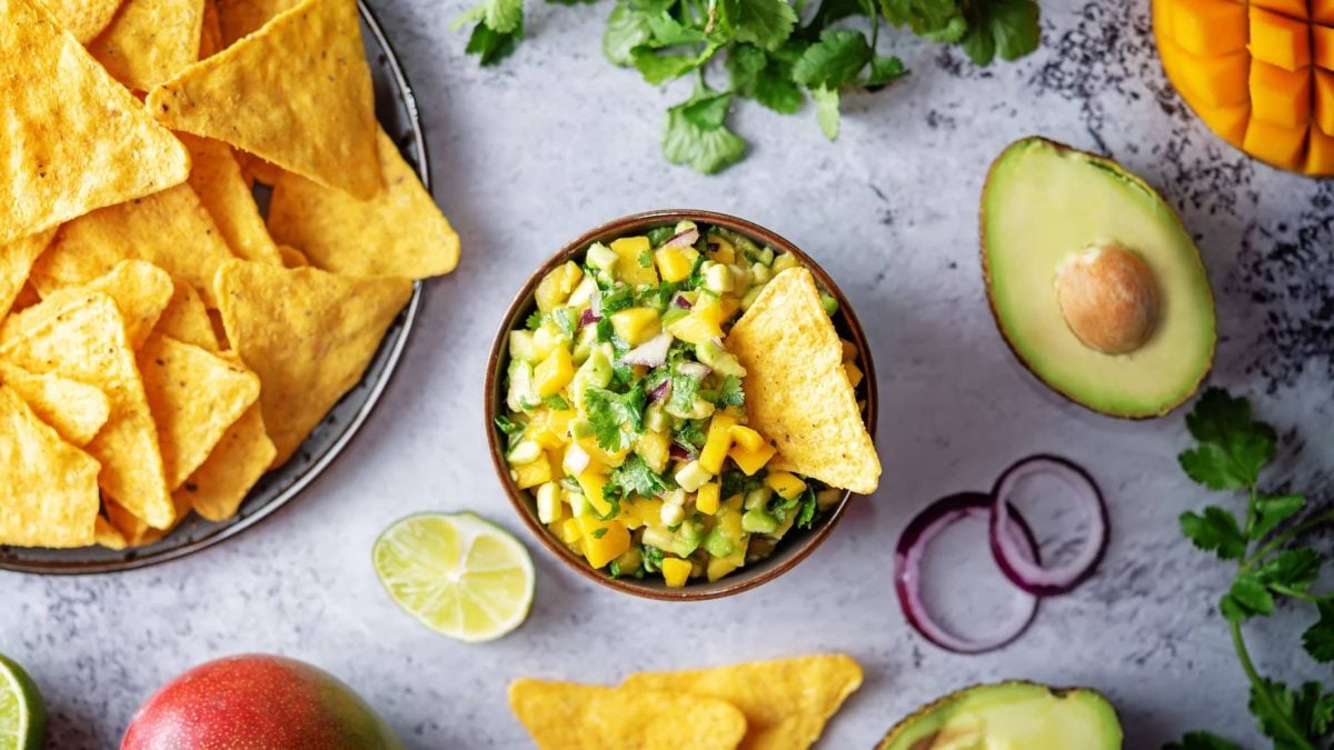 Eine Schüssel Mango-Avocado-Salsa auf grauem Beton in der Draufsicht, daneben liegen Nachos und Zutaten wir Avocado, Mango und Kräutern.