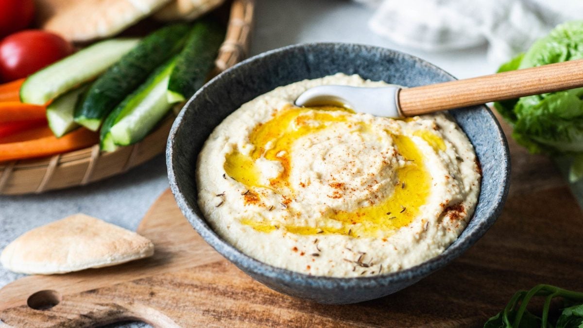 Eine Schale Mangold-Hummus mit Öl und einem Holzlöffe, im Hintergrund Germüse.