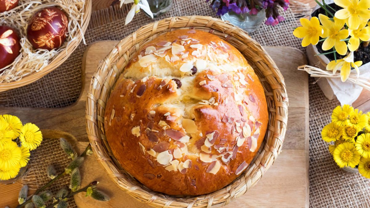 Böhmisches Mazanec Osterbrot mit Rum-Rosinen in Osterkorb auf Holzbrett und Holztisch. Gelbe Blumen und bemalte Eier in Osternest als Deko im Bild. Aufnahme aus der Vogelperspektive.