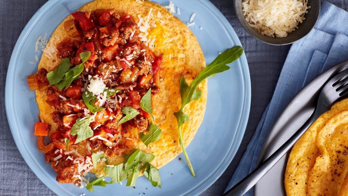 Ein Omelette mit Bolognese, Rucola und Parmesan auf einem blauen Teller in der Draufsicht. Daneben Besteck und eine Schüssel Parmesan.
