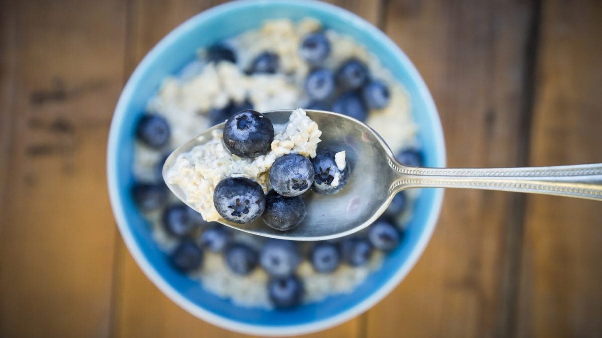 Aus der Vogelperspektive: Ein Löffel mit Overnight Oats mit Blaubeeren. wird über eine Schüssel mit der Frühstücksmahlzeit gehalten.