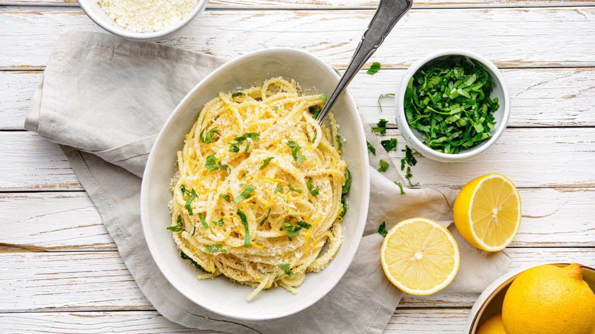 Pasta al Limone in einer weißen Schale. Daneben verschiedene Zutaten. Das Ganze auf einem hölzernen Untergrund.