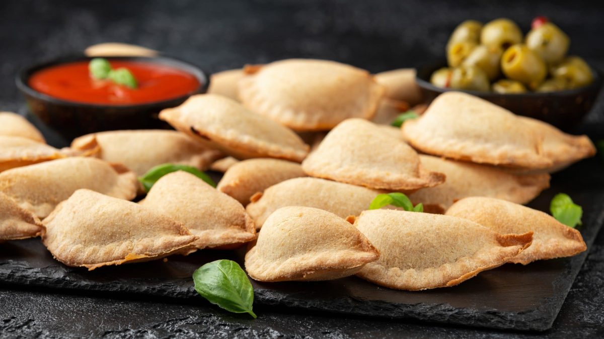 Pizza fritta napoletana auf Schieferplatte. Schwarze Schale mit Tomatensauce und eine weitere mit grünen Oliven im Hintergrund. Basilikumblätter als Dekoration. Frontalansicht.