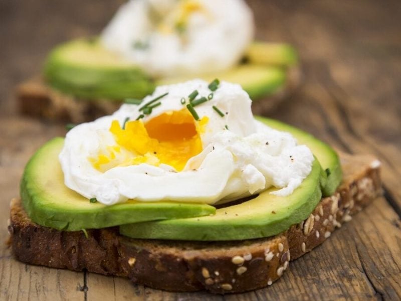 Auf einem hölzernen Untergrund ist im Vordergrund ein Pochiertes Ei auf Avocado-Vollkornbrot. Im Hintergrund ist ein weiteres zu erkennen. Allerdings nur unscharf.