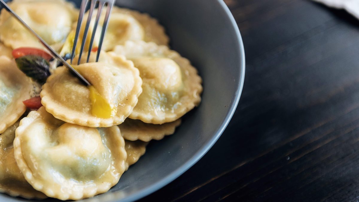 Ravioli in einer grauen Schale und eine Gabel, die in einen hineinsticht. alle spräsentiert auf einem schwarzem Tisch.