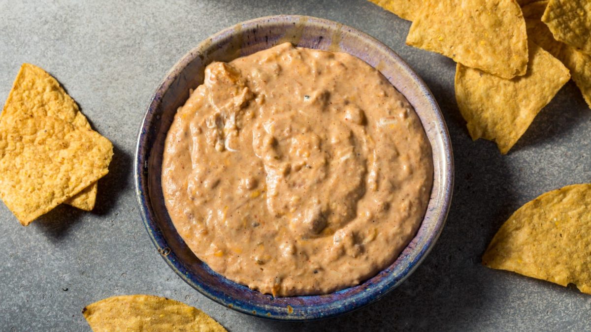 Refried Baked Beans Bohnendip in grauer Schüssel auf grauem Untergrund. Nachos als Dekoration im Bild verteilt. Aufnahme aus der Vogelperspektive.