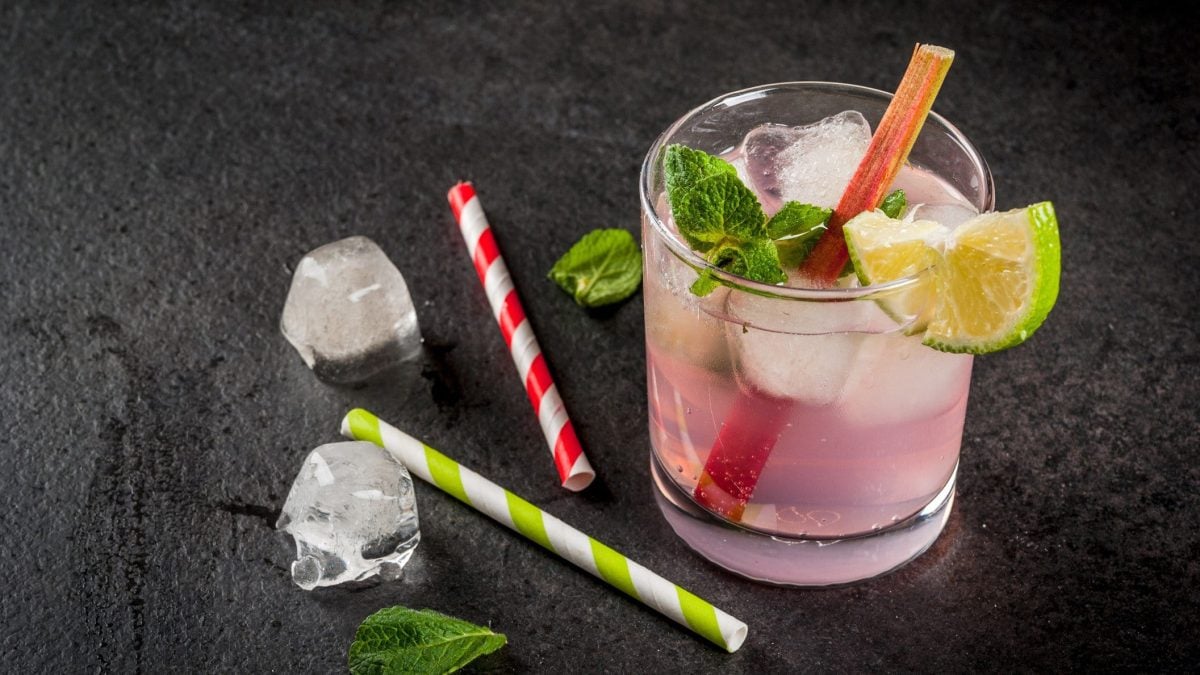 Ein Glas Rhabarber-Spritz mit Minze, Limette und einem Rhabarberstiel, daneben liegen Eiswürfel und Strohhalme aus Papier.