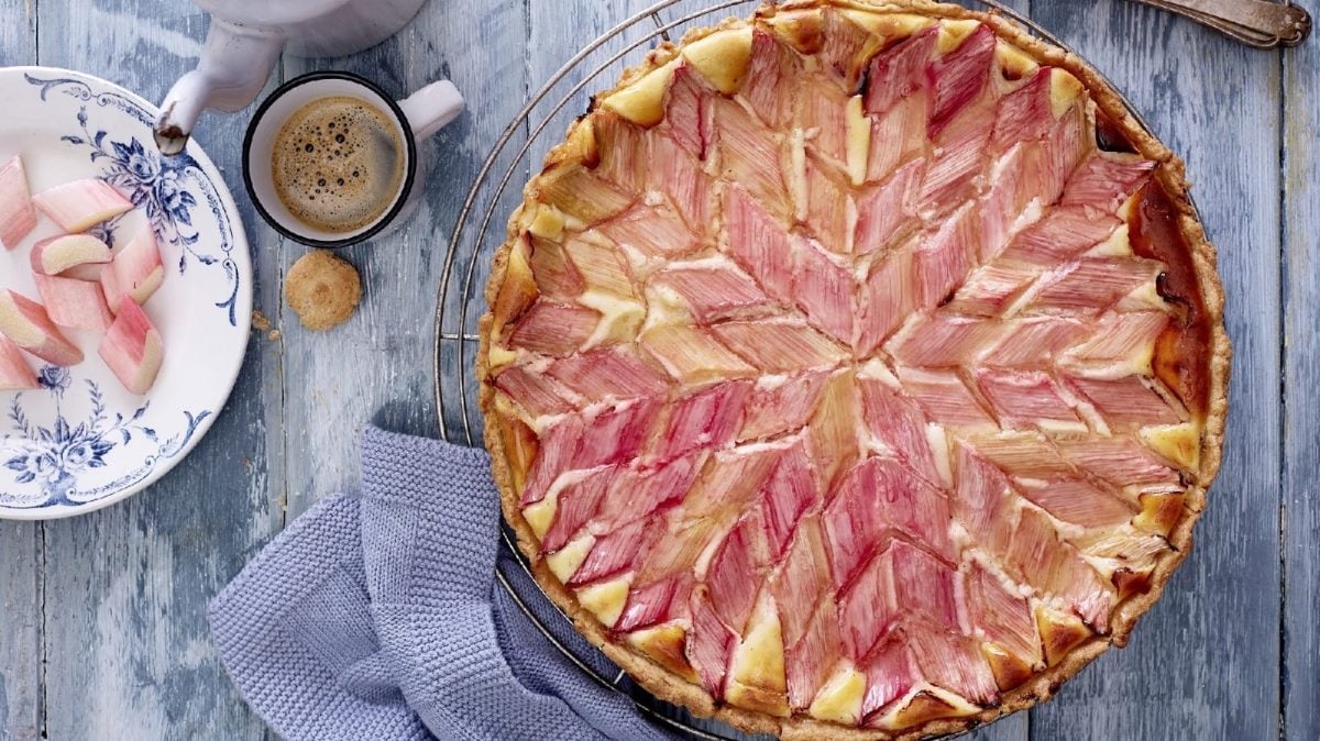 Eine Mascarpone-Rhabarber-Tarte steht auf einem silbernen Gitter auf einem blauen Untergrund. Links neben der Tarte steht eine kleine Schale mit weiteren Rhabarberstückchen und eine Tasse mit Kaffee.