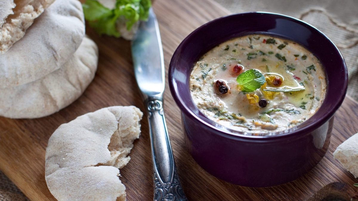 Aus der Vogelperspektive: In eriner kleinen Schlüssel ist ein gebackener Ricotta mit Kräutern. Daneben liegen zwei Flasenbrote, eine Gabel sowie Basilikum-Blätter.