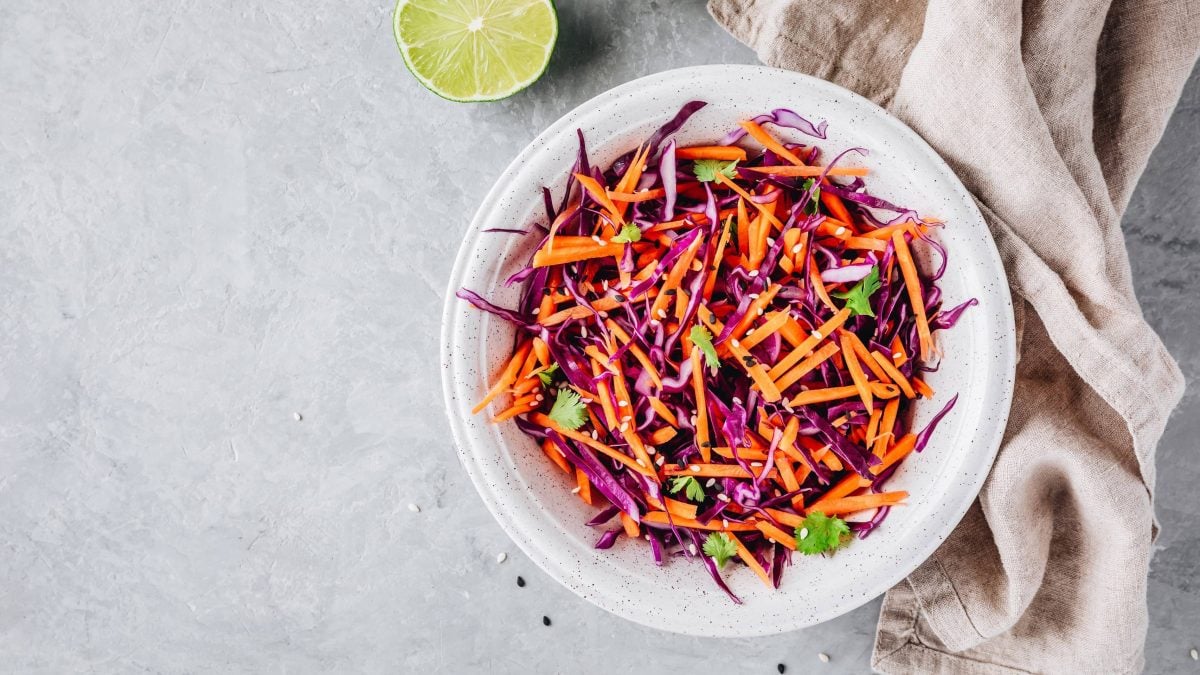 Eine Schüssel Rotkohl-Möhrensalat mit Sesam und Petersilie in der Draufsicht, daneben liegen ein Geschirrtuch und eine halbe Limette.