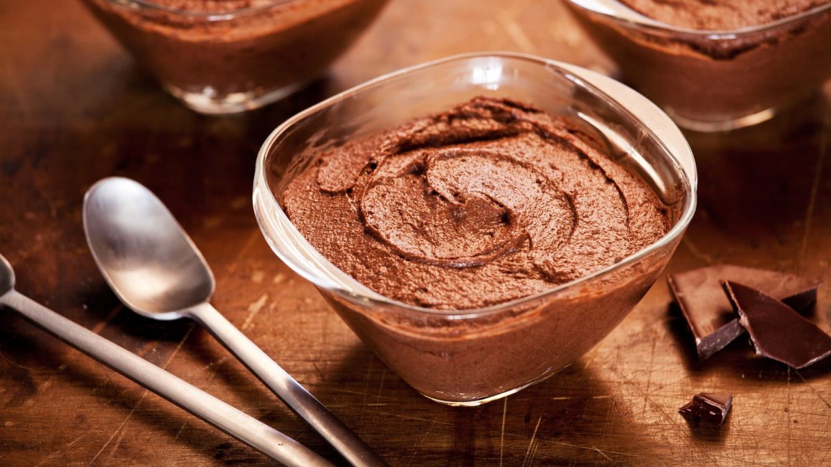 Schnelle Mousse au Chocolat in Glasschale auf braunem Holztisch. Im Hintergrund zwei weitere angedeutet. Links Teelöffel, rechts Schokoladenstücke. Draufsicht.