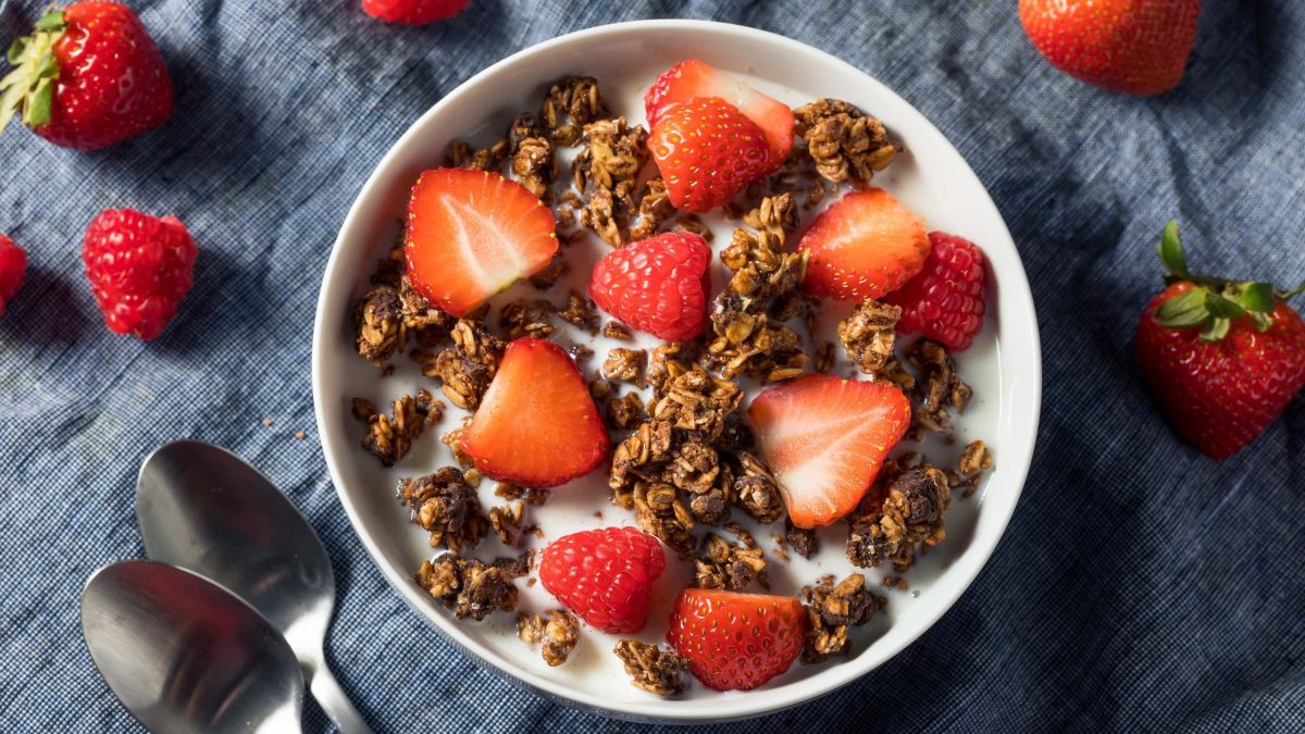 Knuspriges selbst gemachtes Schoko-Granola in einer Schale mit frischen Erdbeeren und Milch, serviert auf einem dunklen Tuch.