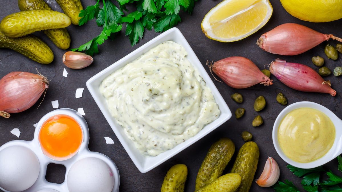 Selbst gemachte Remoulade in einer kleinen Schale. Daneben verschiedene Zutaten auf einem dunklen Untergrund.