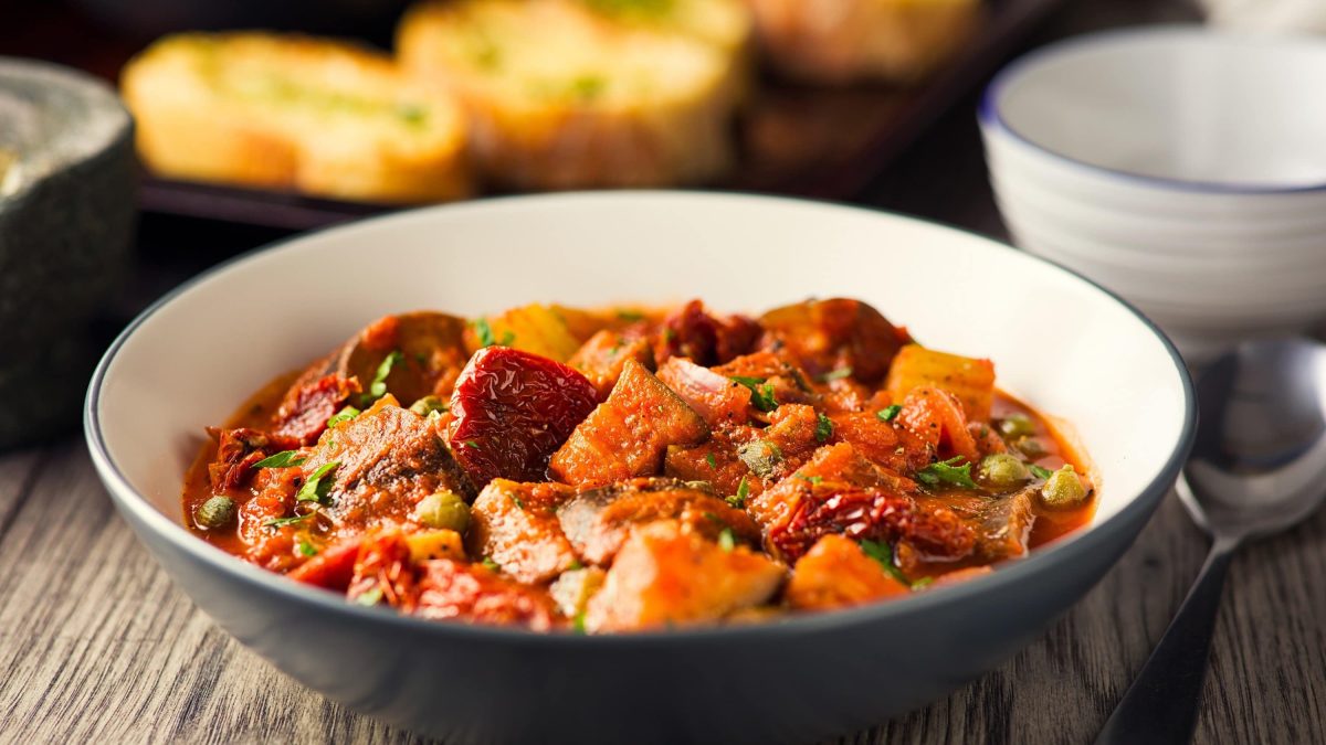 Sizilianische Caponata mit Aubergine, getrockneten Tomaten, Paprika und frischer Petersilie in einer großen Schale auf rustikalem Holztisch.